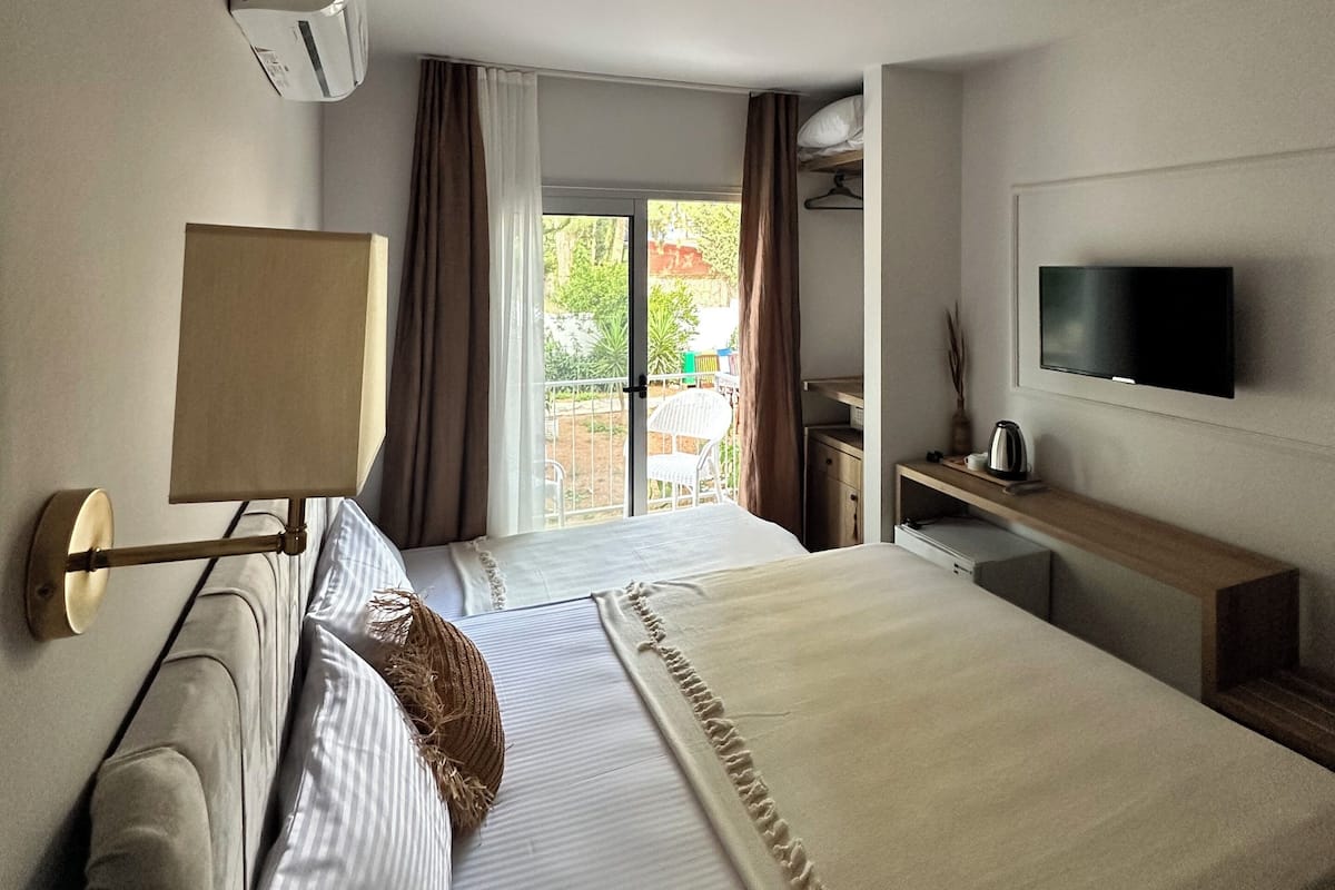 Un cuarto de hotel con una cama grande, un televisor montado en la pared, una mesita de noche con lámpara, y vistas al exterior a través de la ventana.
