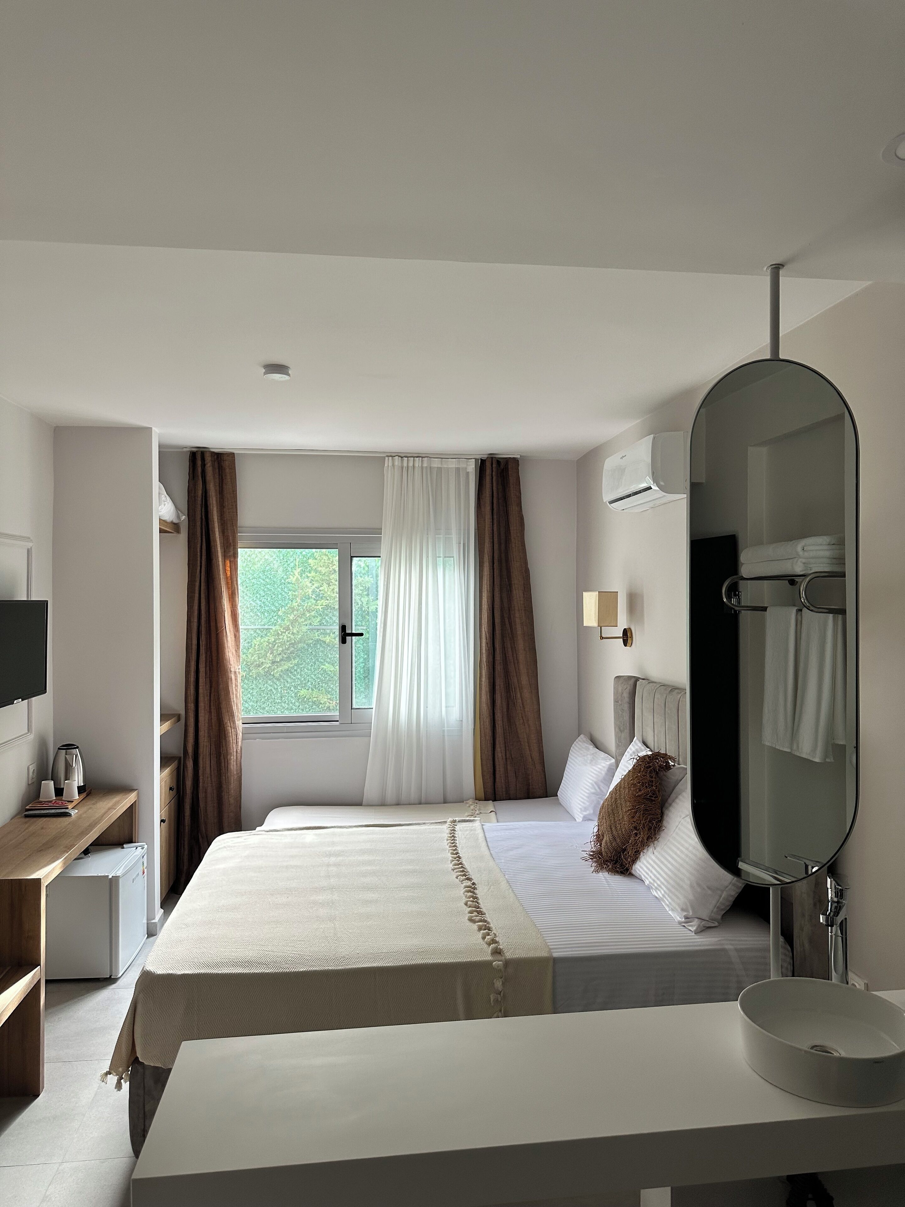 A hotel room with a bed, a bedside table with a kettle, a mirror, and a window with curtains.