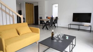 A modern living room with a yellow sofa, a dining area with a table and chairs, a black coffee table with wine glasses and a bottle, and a TV on a stand. - Gîte L'angélique - Fleurs des Pyrénées - Vue Hautacam (Argelès-Gazost)