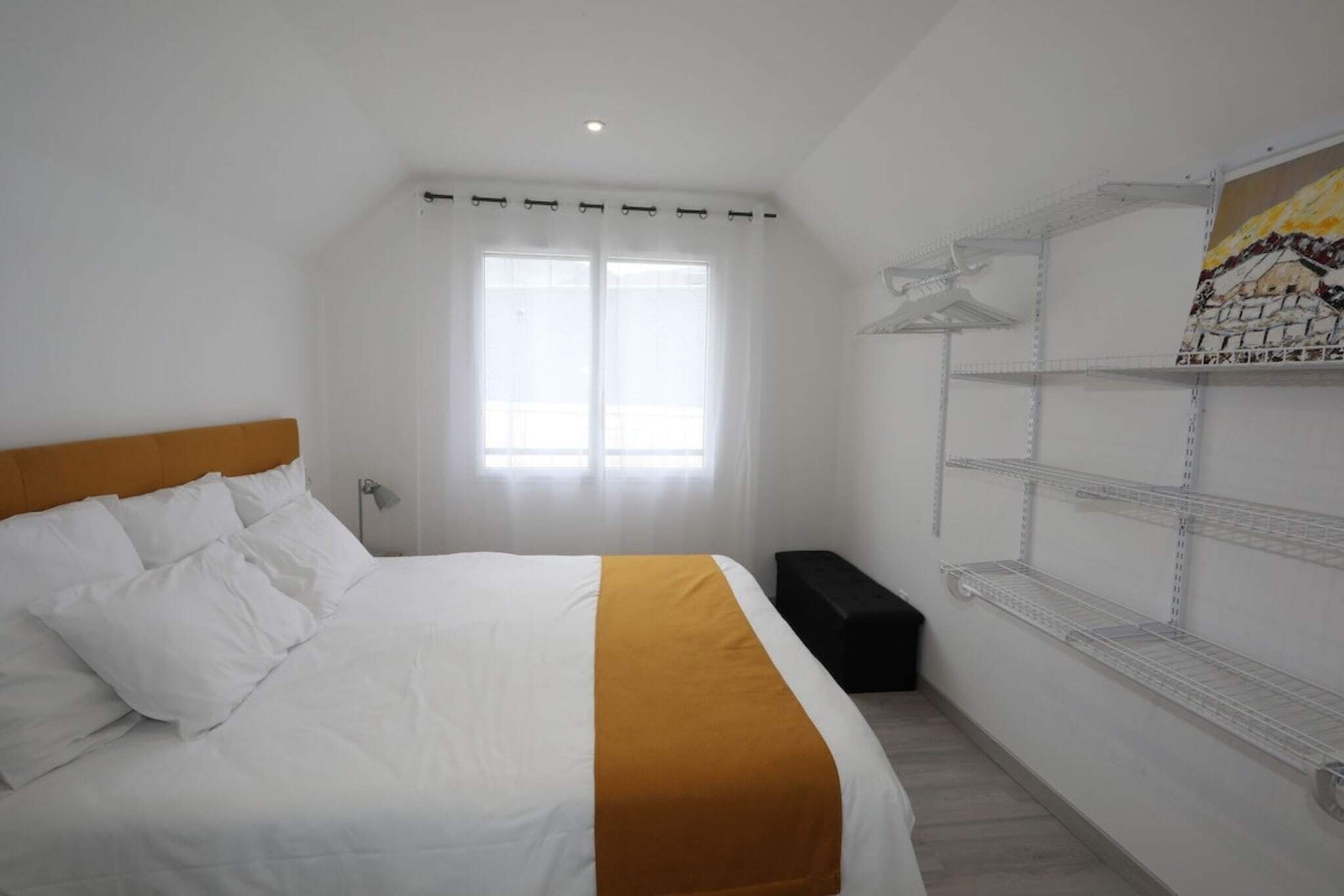 A bedroom with a bed, a window with curtains, a bedside table, and a wall-mounted shelf.