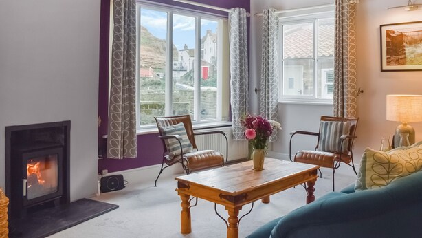 Een woonkamer met een open haard, twee ramen met gordijnen met een patroon, een houten tafel met een bloemenvaas, en een blauwe bank.