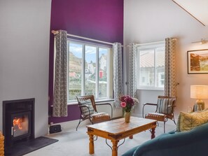Een woonkamer met een open haard, twee ramen met gordijnen met een patroon, een houten tafel met een bloemenvaas, en een blauwe bank.