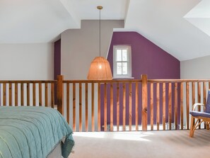 Een slaapkamer met een schuine balkenkap, houten leuning, bed met een blauwe deken, een rieten stoel en een hangende lamp.