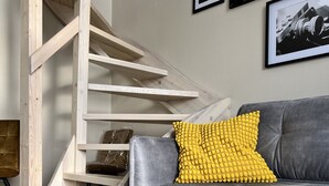 Sala moderna com sofá cinza, almofada amarela e escada de madeira.