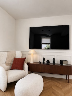 A modern living room with a white sofa, a red pillow, a round ottoman, a wooden side table with a lamp, and a flat-screen TV mounted on the wall.