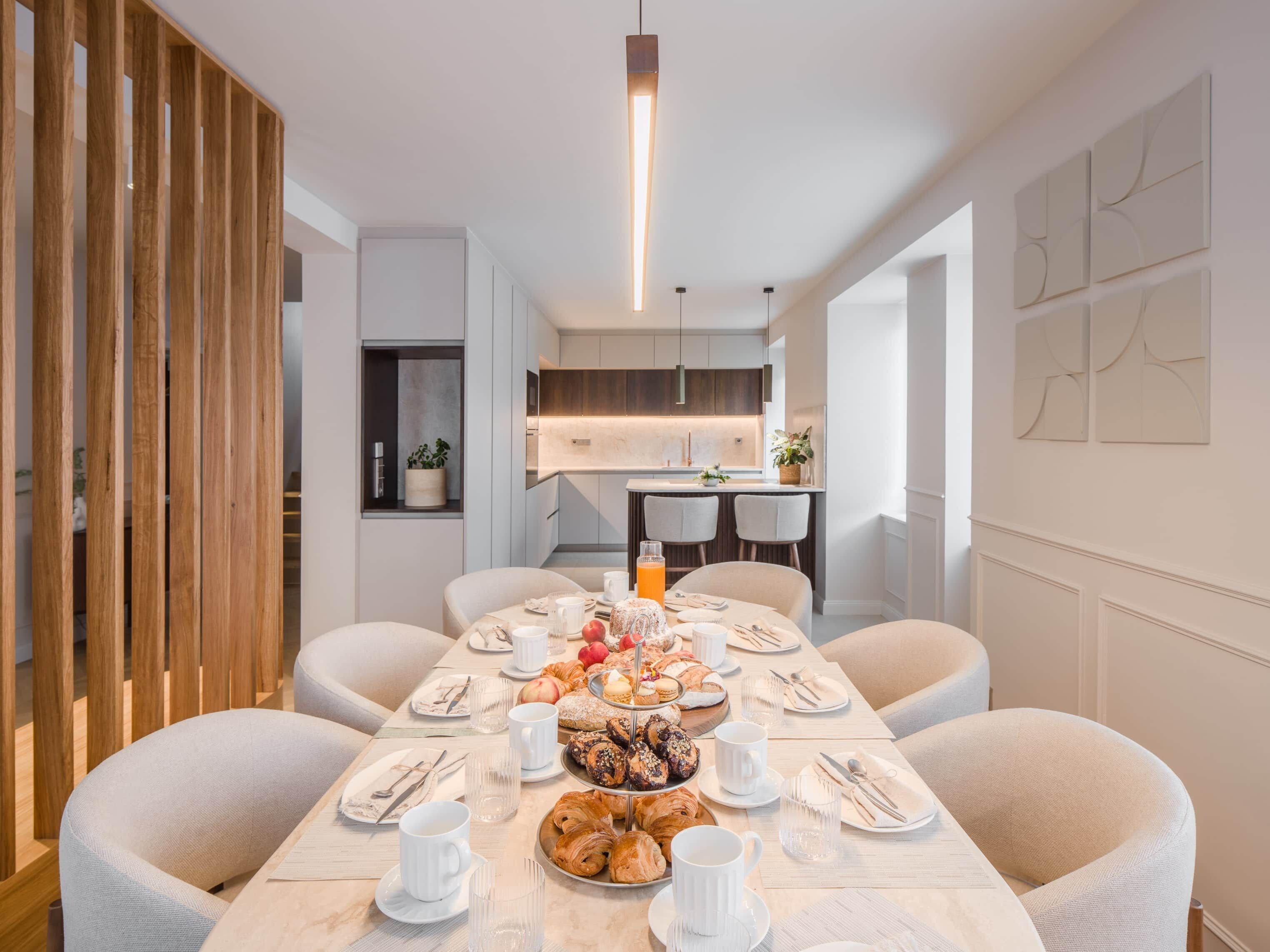 Un coin repas avec une longue table dressée pour le petit-déjeuner, une cuisine en arrière-plan et un aménagement intérieur moderne.