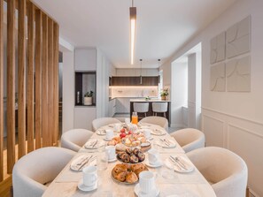 A dining area with a long table set for breakfast, a kitchen in the background, and a modern interior design.
