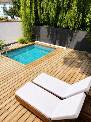 Une terrasse en bois avec une piscine, entourée de verdure et d’une clôture sombre.