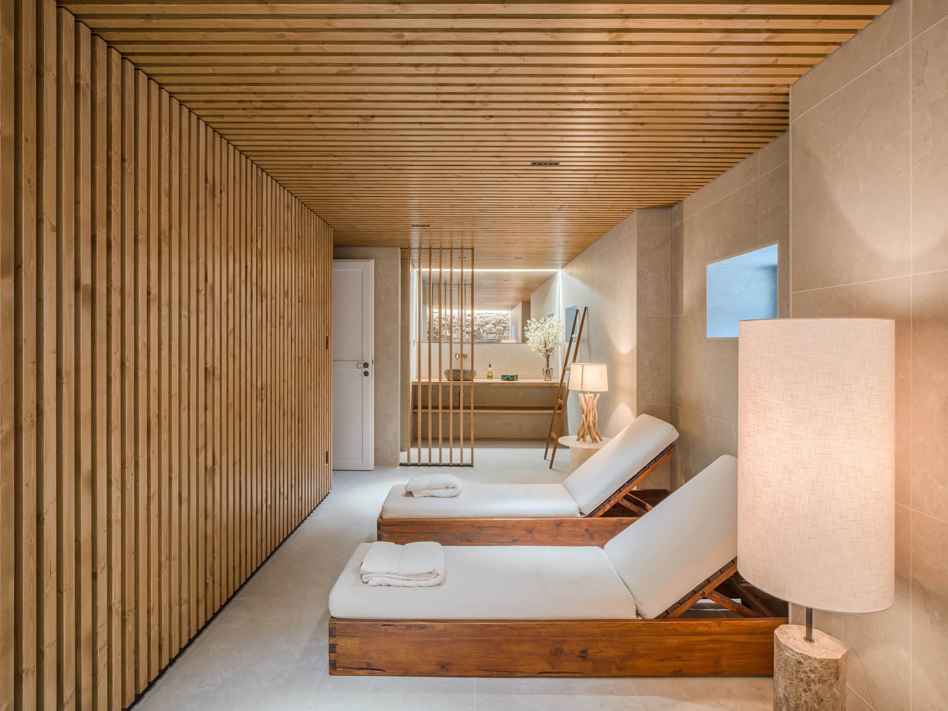 Ein Spa-Raum mit Holzlatten an den Wänden, zwei Holzliegen mit Handtüchern, ein Steinfußboden, eine Steintanne und Blick auf eine Steinwand und ein Fenster.