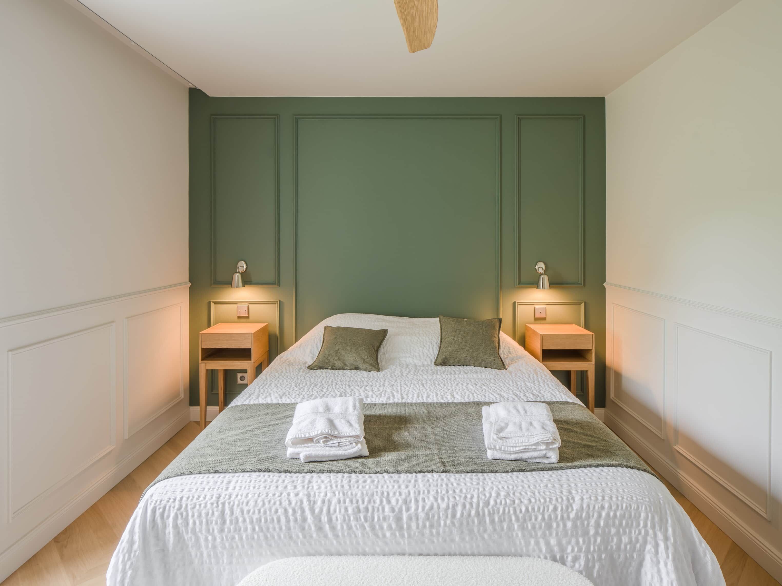 A neatly made bed with pillows and folded towels, two bedside tables with lamps, and a green wall paneling.