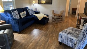 Una sala de estar con un sofá azul, un sillón estampado, piso de madera, una lámpara y un cuadro en la pared.