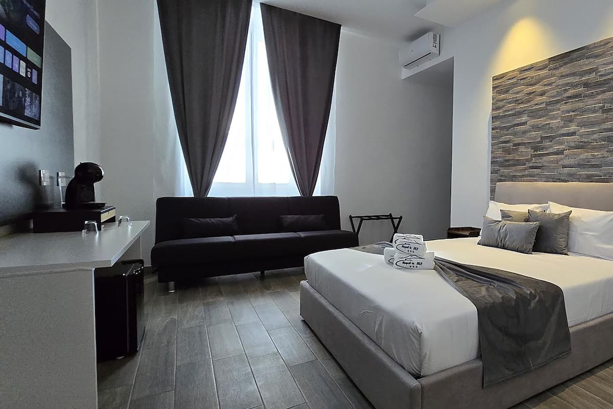 A modern hotel room with a large bed, a sofa, a desk, and a stone-textured wall.