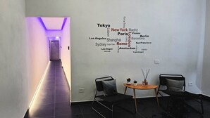 Un pasillo moderno con una nube de palabras en la pared que muestra nombres de ciudades. Hay dos sillones y una pequeña mesa con una vela y una planta.
