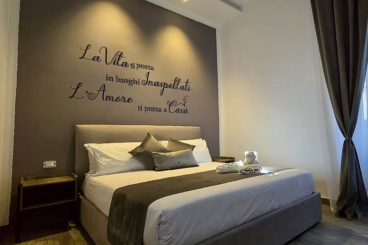 A modern hotel room with a large bed, a bedside table, a nightstand, and a wall with a decorative phrase.