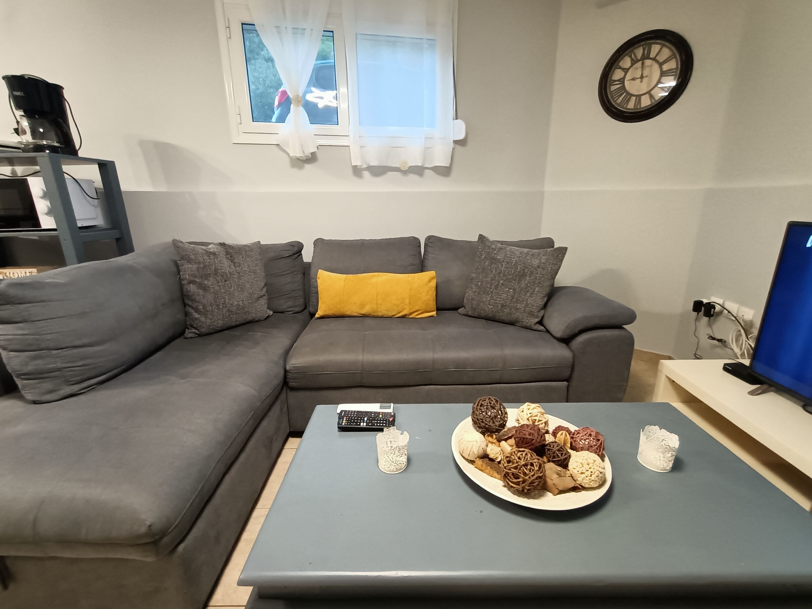 A living room with a grey sectional sofa, a coffee table with decorative items, a clock on the wall, and a television on a stand.