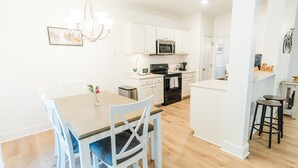 A modern kitchen with white cabinetry, a central island, and a dining area with a wooden table and white chairs. - Claire's Charming Home near RDU and RTP (Raleigh)