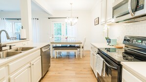 A modern kitchen with white cabinetry, stainless steel appliances, a wooden dining table with chairs, and a chandelier. - Claire's Charming Home near RDU and RTP (Raleigh)