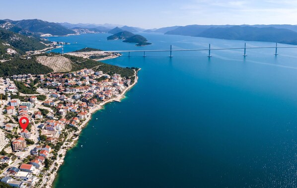 An aerial view of a coastal town with a bridge, clear blue sea, and hilly terrain in the background.