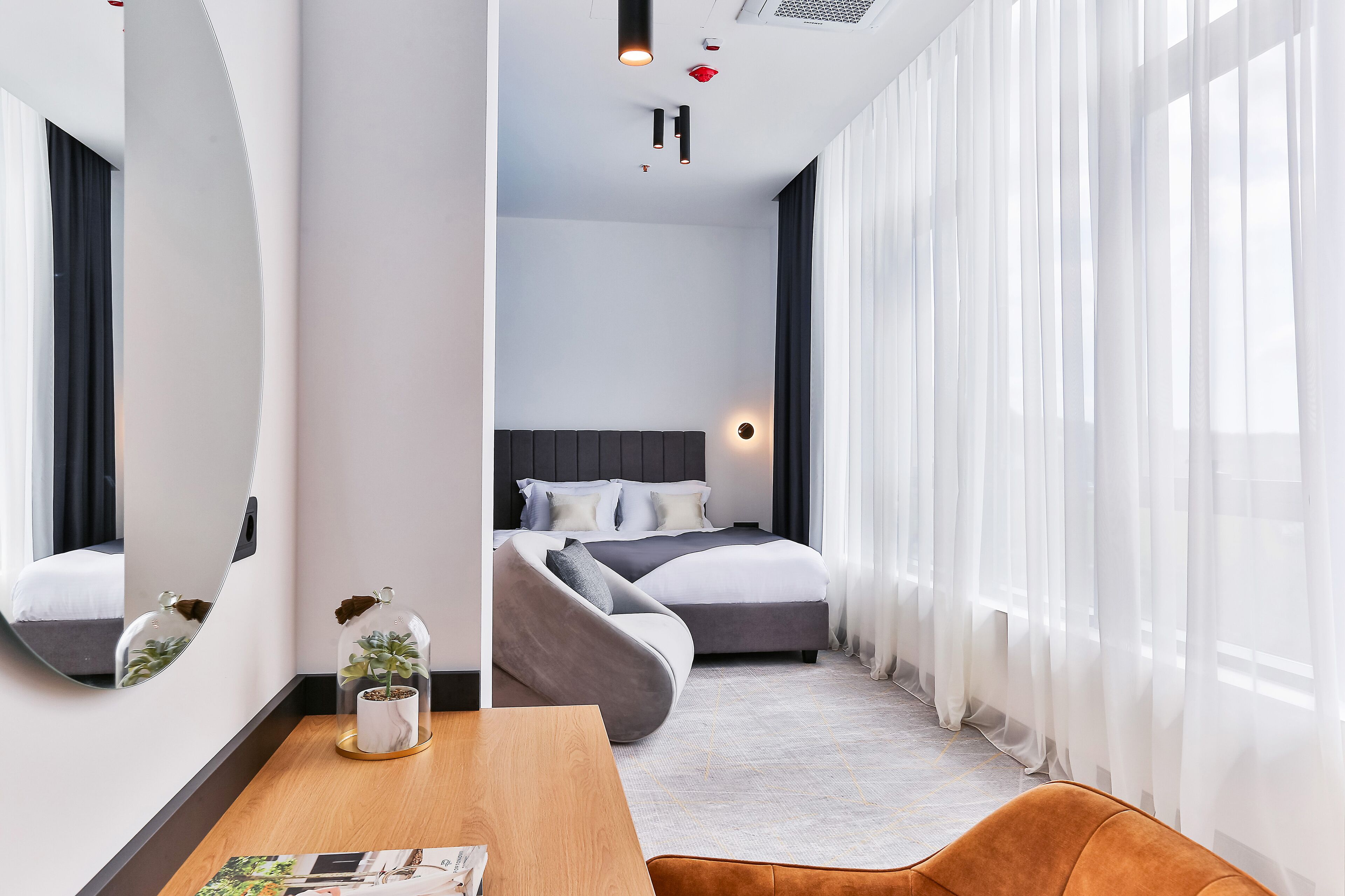 Et moderne hotelværelse med et buet spejl, en seng med gråt sengetøj, et træbord og lette gardiner.