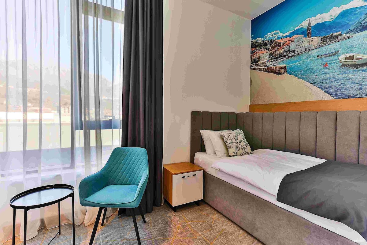 A hotel room with a bed, a chair, a small table, and a painting of a coastal town on the wall.