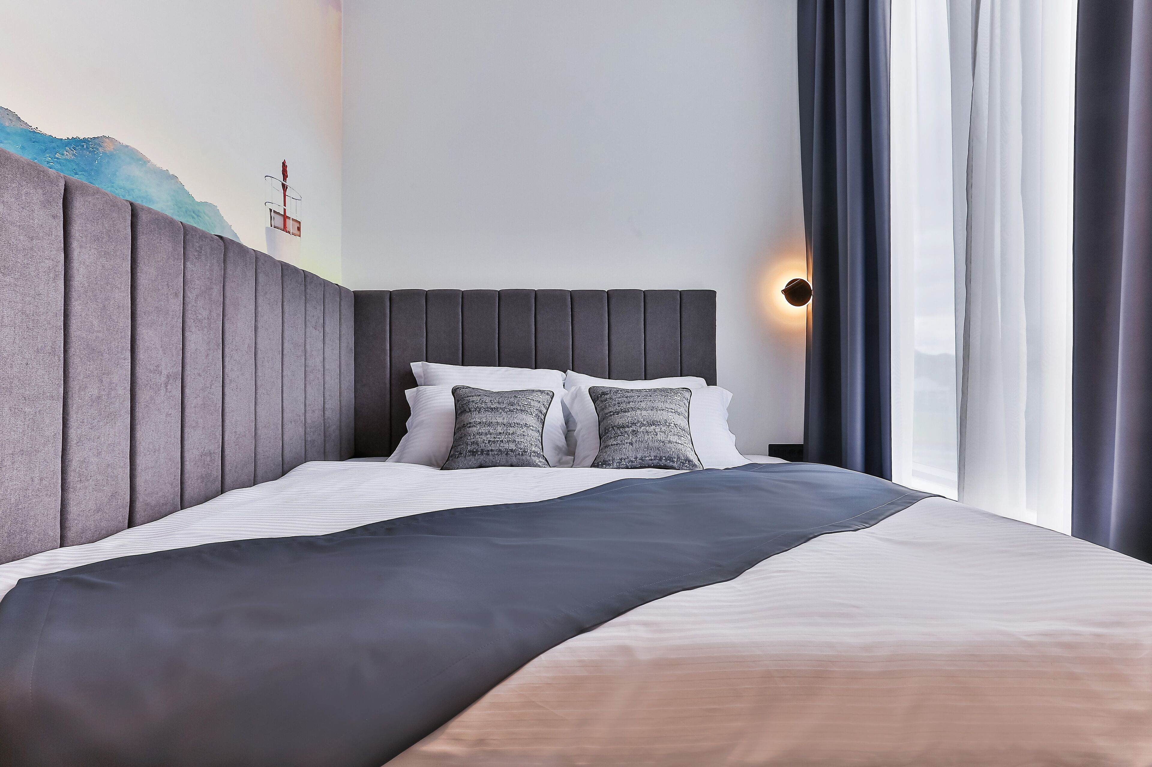 Una habitación de hotel moderna con una cama grande, cabecera gris, ropa de cama blanca y una lámpara de pared.