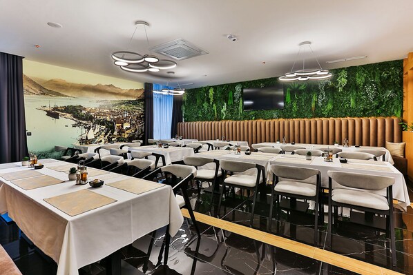 A restaurant with multiple tables and chairs, a mural of a coastal town, and a green wall with plants.