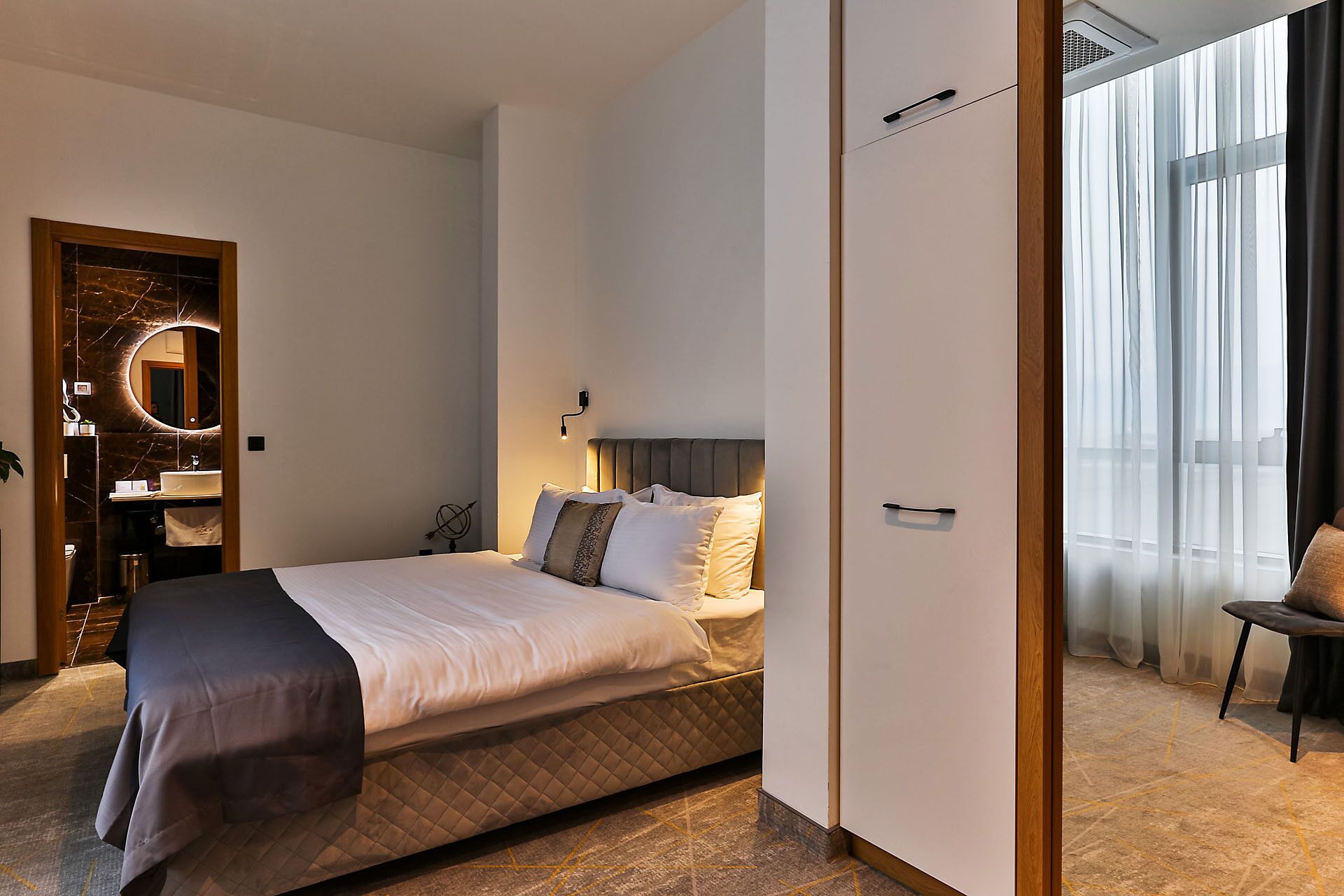 A modern hotel room with a bed, a bedside table, a mirror, and a view of the outdoors.