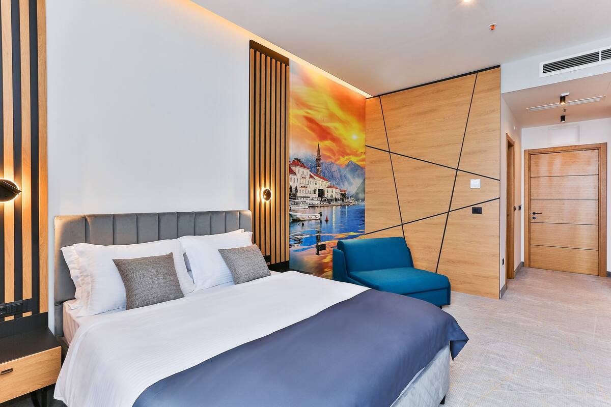 A modern hotel room with a large bed, a blue armchair, and a wall mural depicting a cityscape.