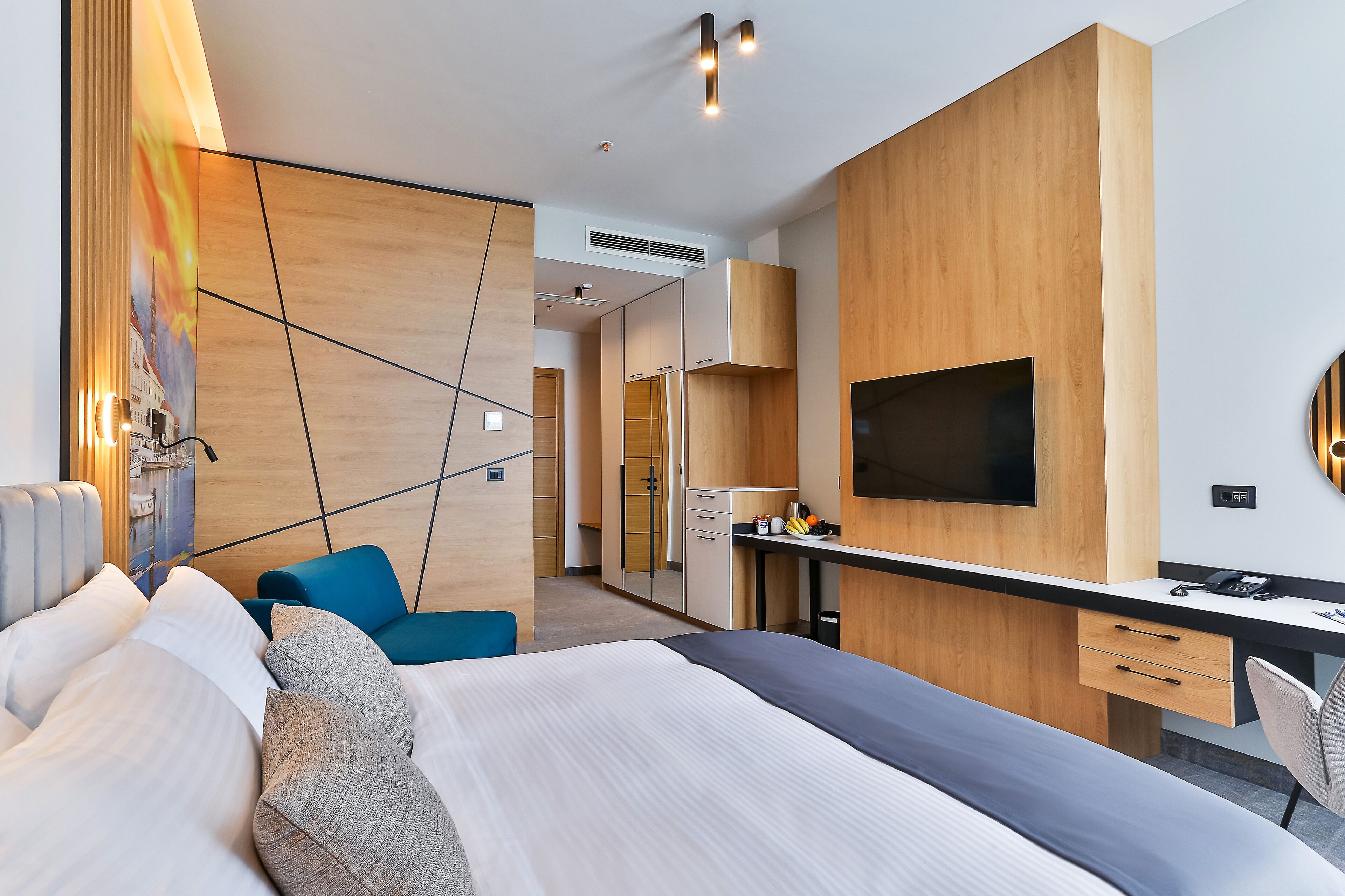 Una habitación de hotel moderna con una cama grande, un escritorio con televisor de pantalla plana, una silla azul y un armario de madera.