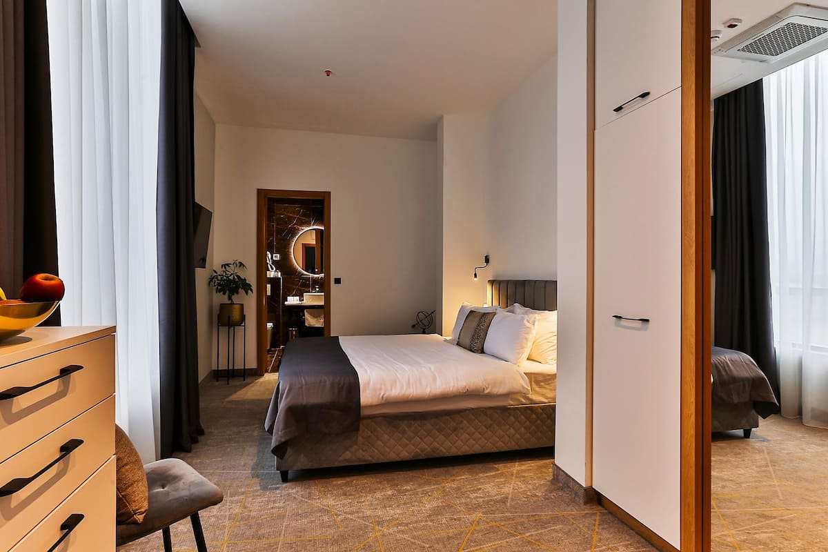 A modern hotel room with a bed, a desk, and a dresser. There is a bathroom visible through an open door.