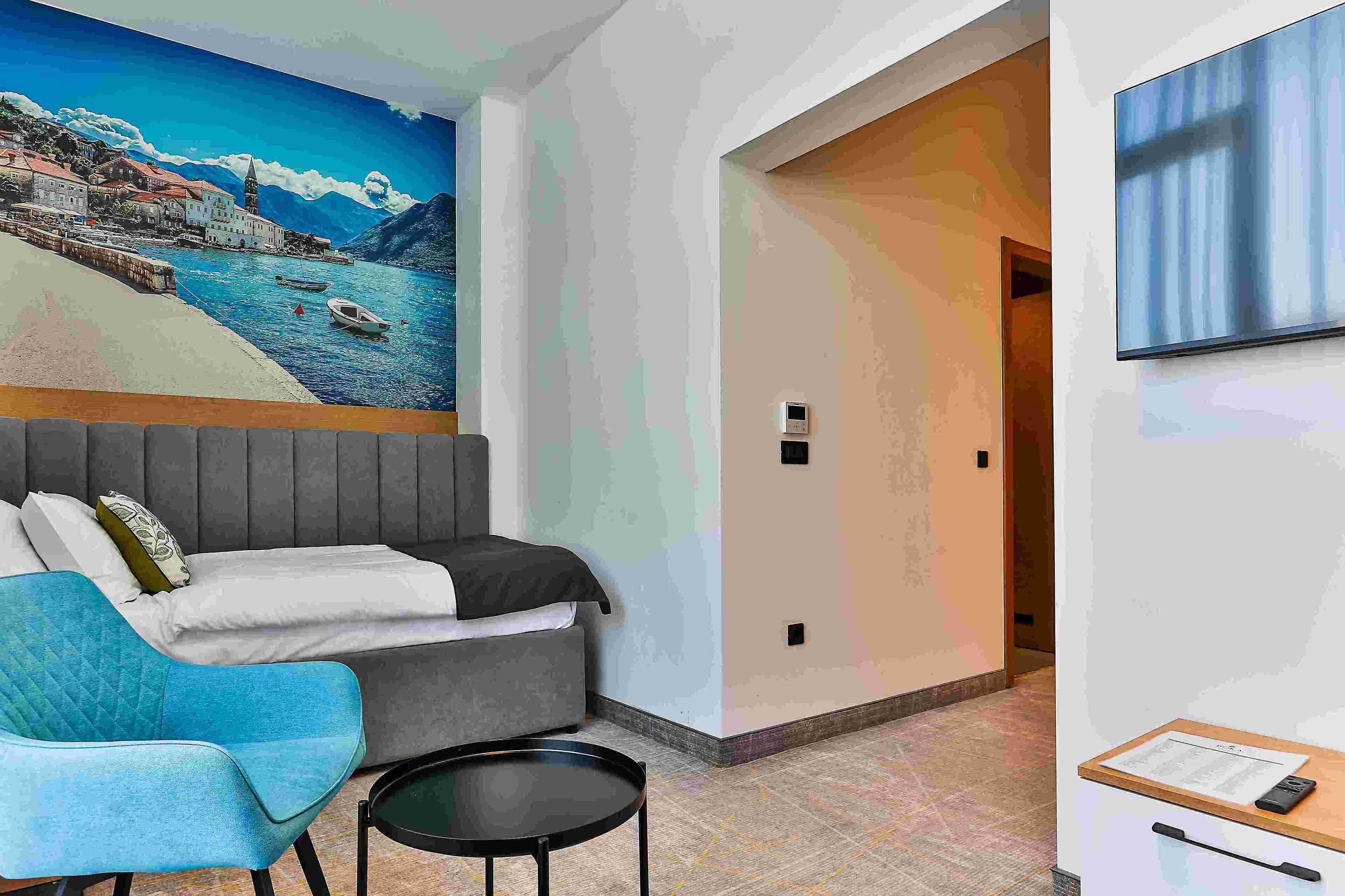 A hotel room with a bed, a blue armchair, a small table, a mural of a coastal scene, and a TV on the wall.