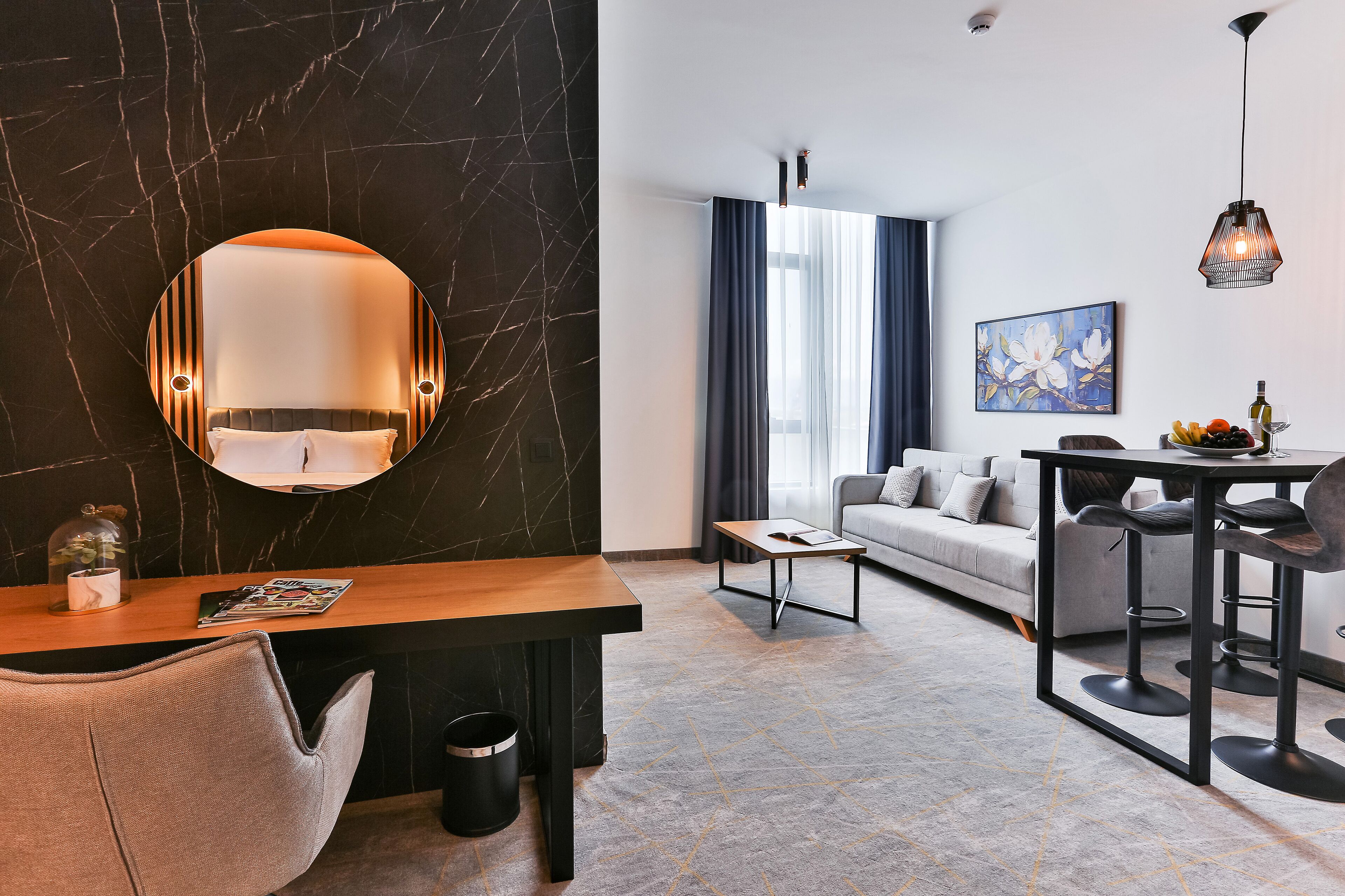 Una habitación de hotel moderna con una pared decorada con mármol, un espejo redondo, un escritorio de madera, un sofá gris, una zona de comedor con una mesa y sillas, y un ventanal con cortinas.
