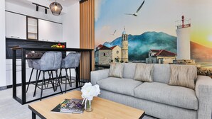 A modern living room with a grey sofa, a wooden coffee table with a magazine and a vase of flowers, a dining area with a black table and grey chairs, and a mural of a coastal town with mountains and a lighthouse. - Hotel Boka (Radanovici)