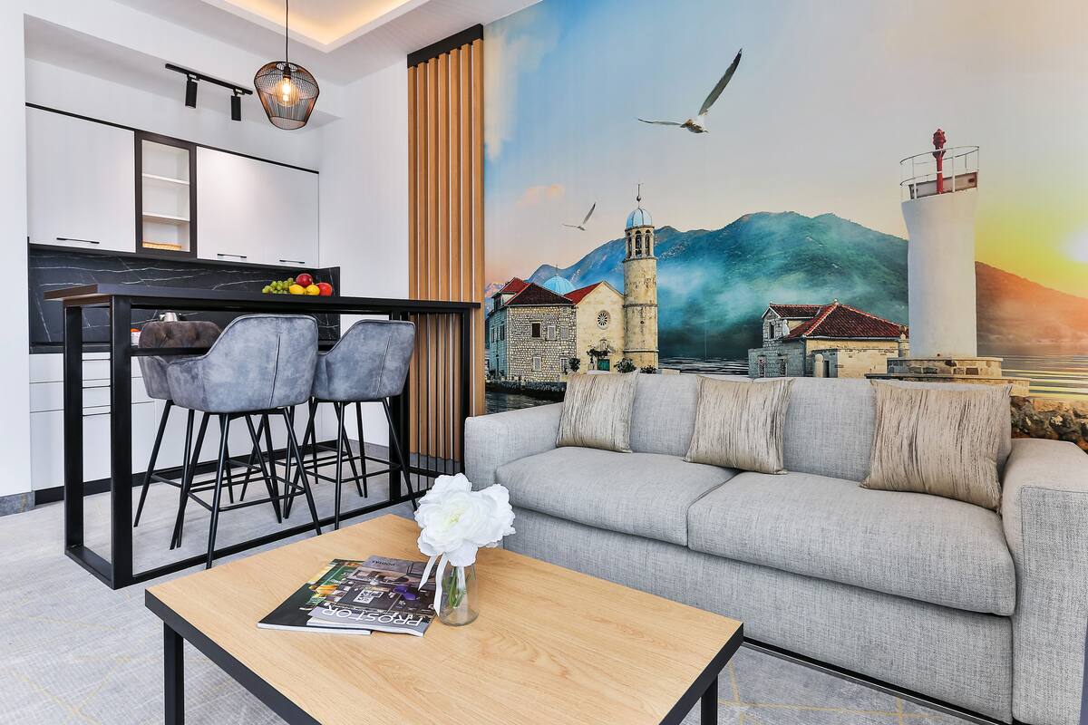 A modern living room with a grey sofa, a wooden coffee table with a magazine and a vase of flowers, a dining area with a black table and grey chairs, and a mural of a coastal town with mountains and a lighthouse.