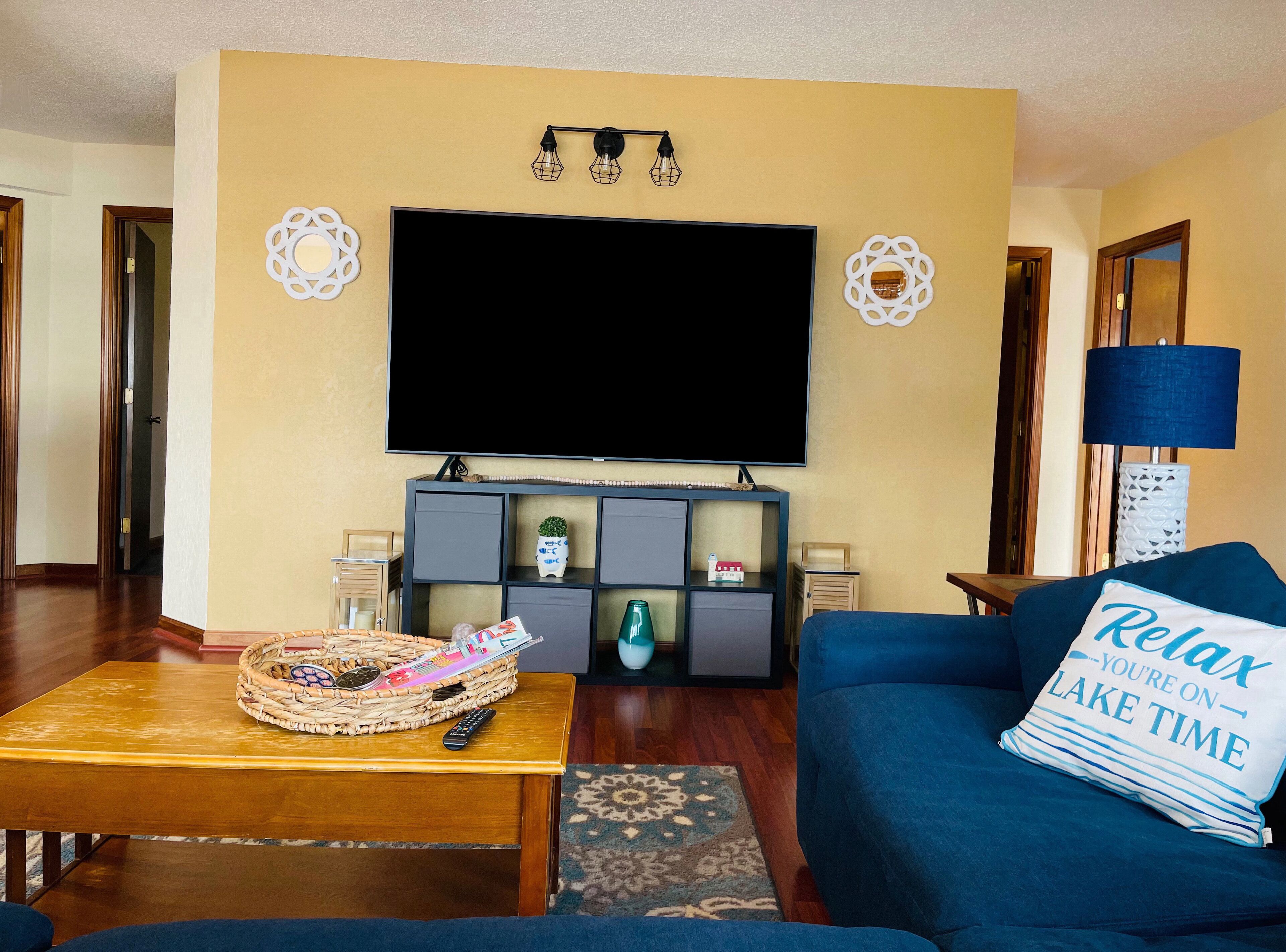 Una sala de estar con un televisor de pantalla plana, un sofá azul con un cojín, una mesa de centro de madera con una cesta y el control remoto, y apliques decorativos en la pared.
