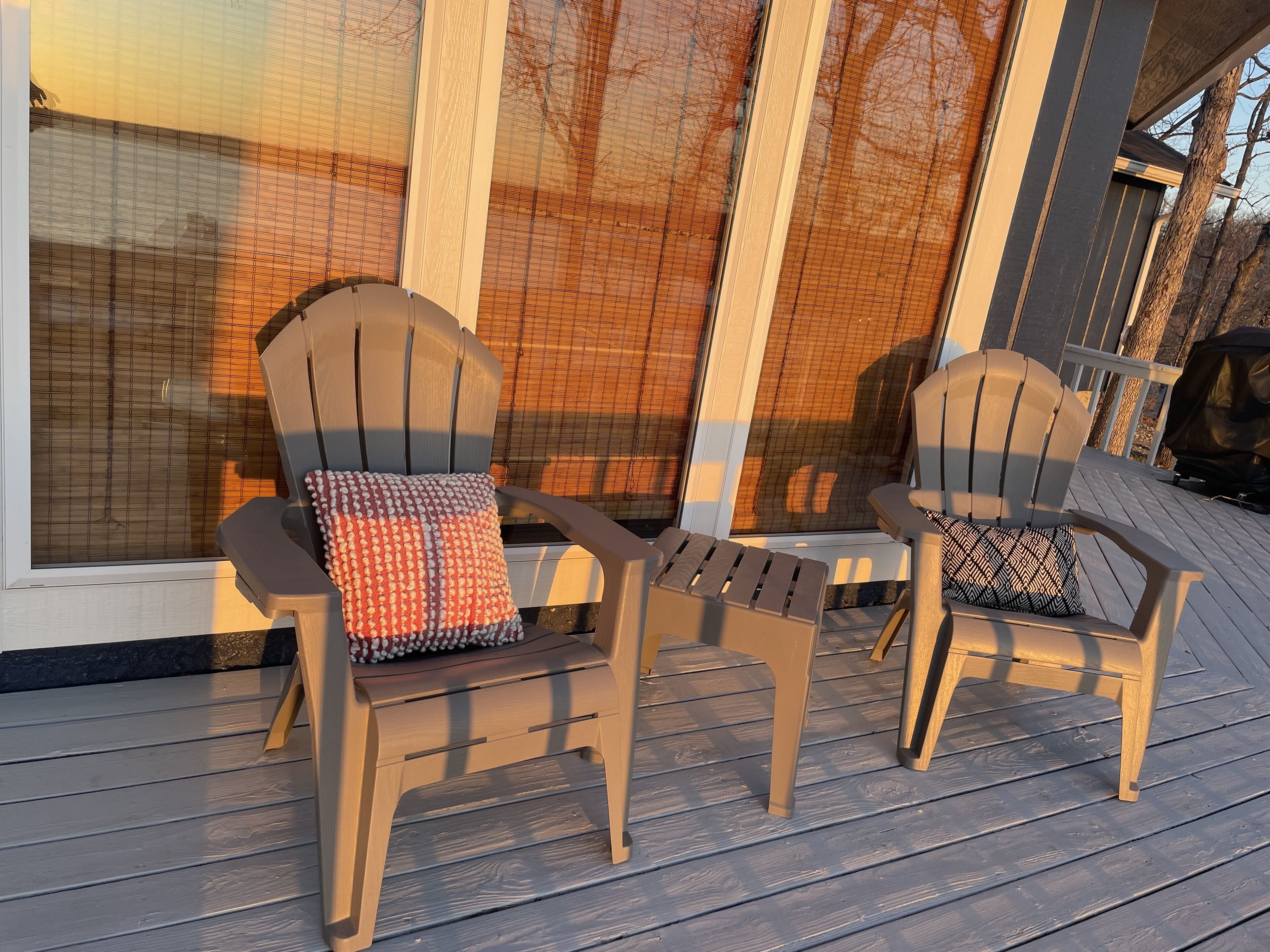 Two Adirondack chairs with patterned cushions on a wooden deck, a large window with a view of trees, and a grill in the background.