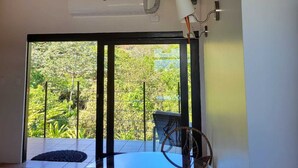 Una habitación con techo de madera, ventilador de techo, ventana grande con vistas a follaje, mesa con sillas y una alfombra pequeña.