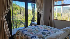 Un dormitorio con cama, cortinas y vistas a la vegetación a través de amplios ventanales.