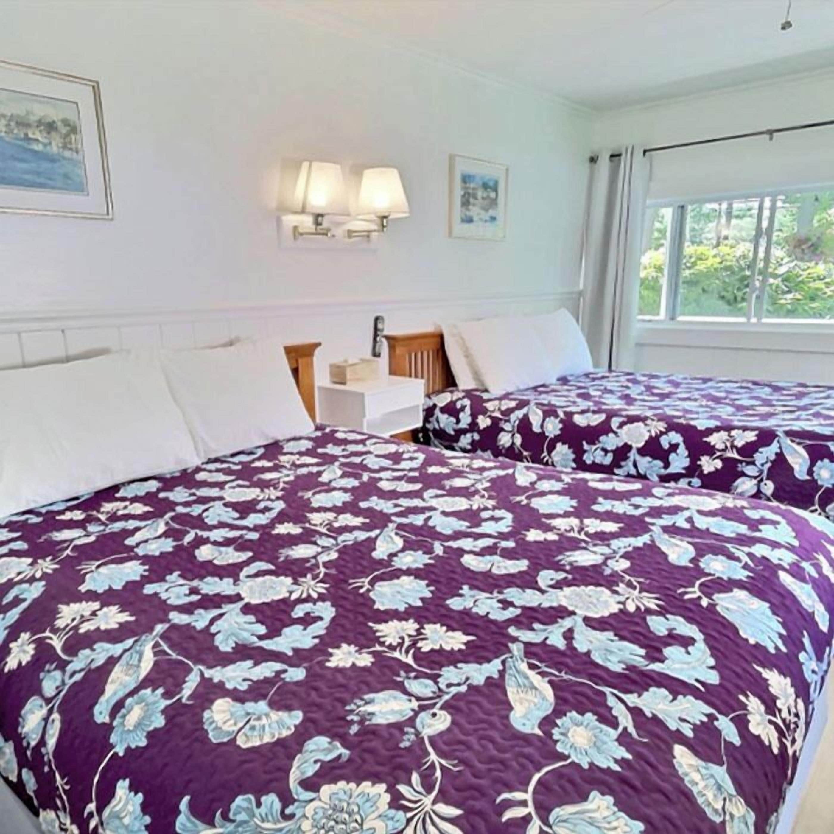 A hotel room with two beds, a nightstand, a lamp, and a window with a view of greenery.