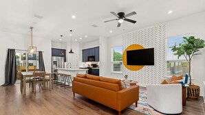 A modern living room with a sofa, dining table, and chairs, a kitchen in the background, and a large TV mounted on the wall.