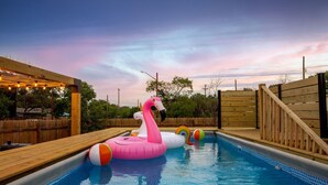 A swimming pool with inflatable flamingo and rainbow floats, surrounded by wooden deck and a wooden fence.
