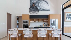 A modern kitchen with a central island, wooden chairs, and a black pendant light.