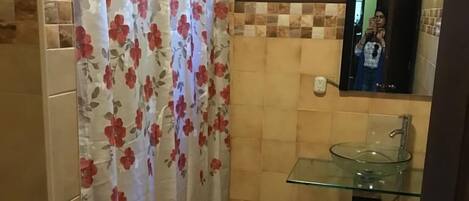 Uma casa de banho com lavatório de vidro, separador de chuveiro com padrão floral e chão em mosaico com design geométrico.