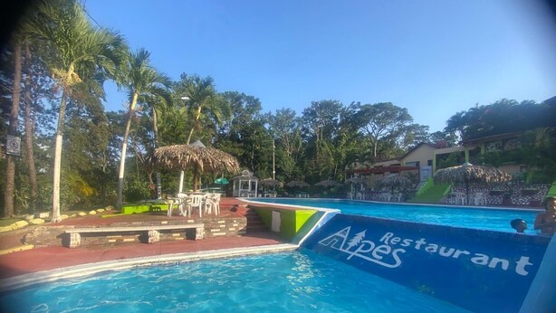 A swimming pool with a restaurant sign, surrounded by palm trees and a gazebo. - Brisas de los Alpes (Jarabacoa)