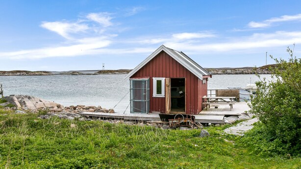 A small, red wooden boathouse with a metal door and windows, situated by a calm body of water with a dock extending into the water.