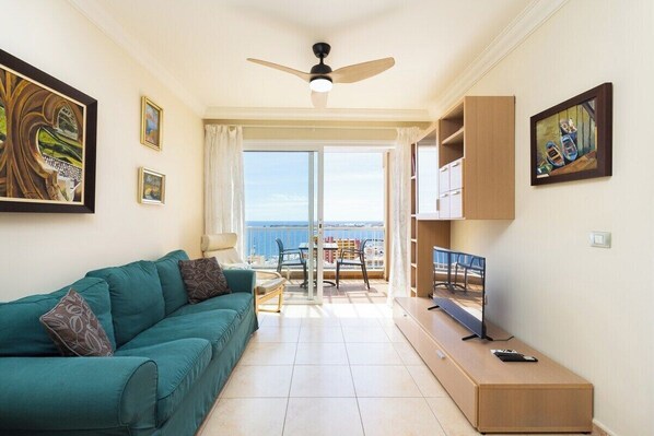 Un salon avec un canapé de couleur turquoise, un ventilateur de plafond, un téléviseur posé sur un meuble en bois et un balcon avec vue.