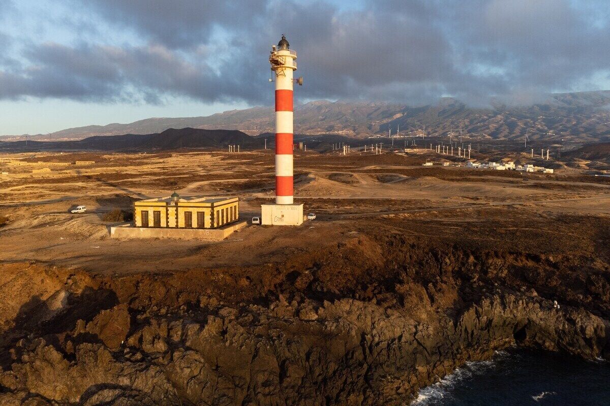Ein Leuchtturm mit einem rot-weiß gestreiften Turm, ein kleines Gebäude mit einer gelben Fassade und eine felsige Küstenlinie mit Bergen im Hintergrund.