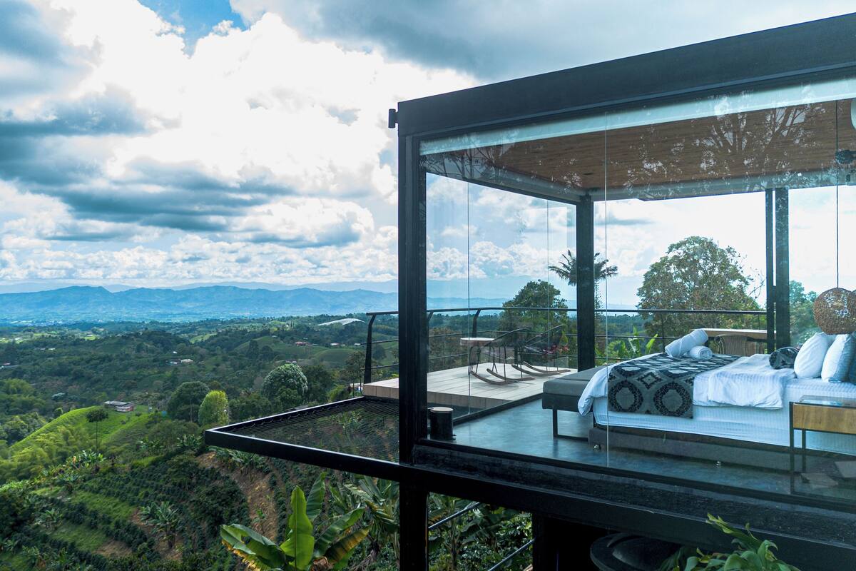 Ein moderner Glasbalkon mit Bett und Sitzgelegenheit mit Blick auf eine üppige Landschaft und entfernte Berge.