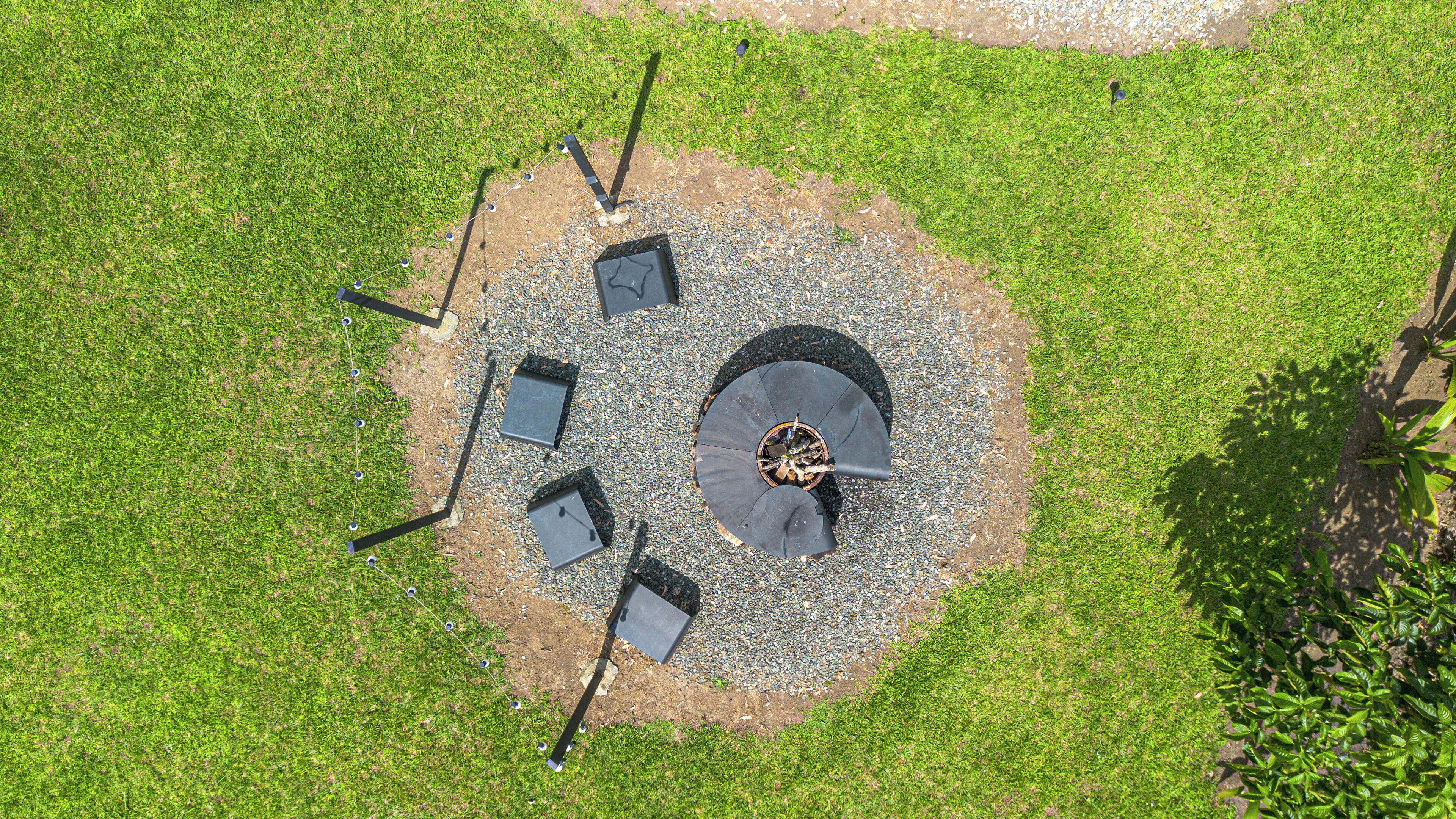 An aerial view of a circular outdoor seating area with a central fire pit, surrounded by a gravel path and bordered by grass.