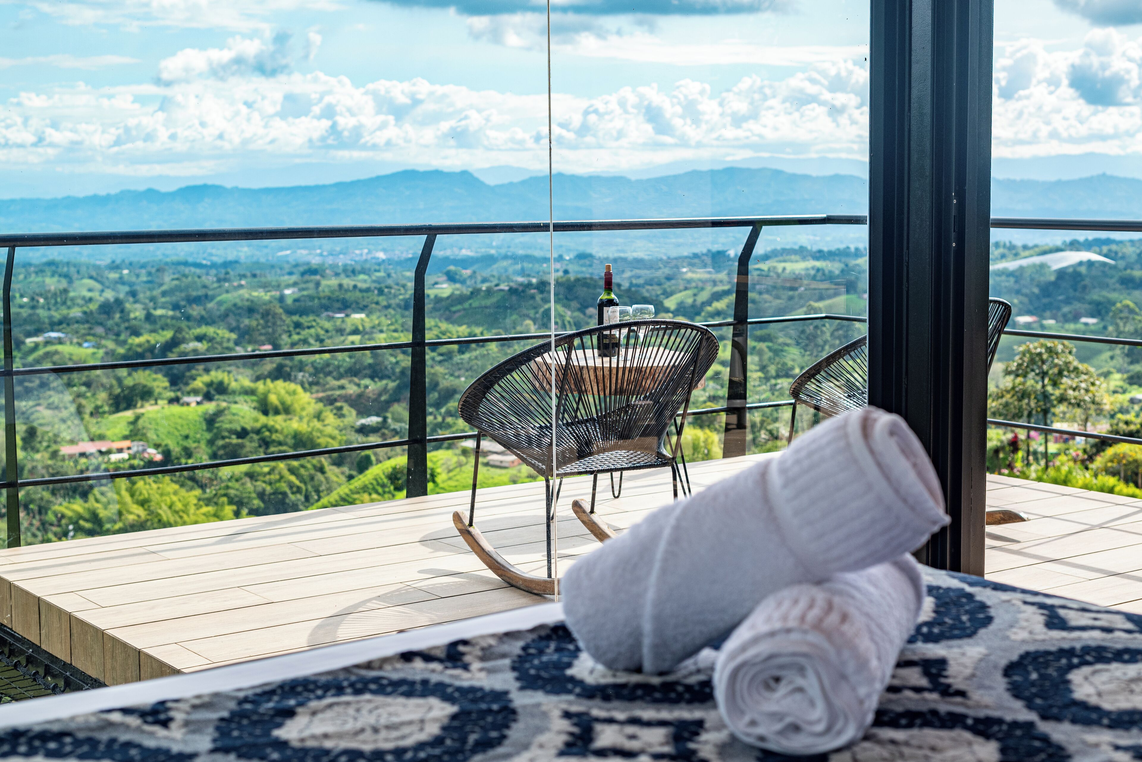 A balcony with a view of a mountainous landscape, a wicker chair, and a rolled-up towel on a patterned rug.